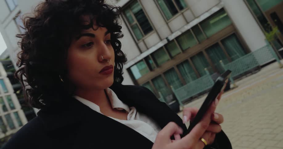A woman with curly dark hair and a nose ring in a black coat uses her smartphone while standing outdoors near a large modern building