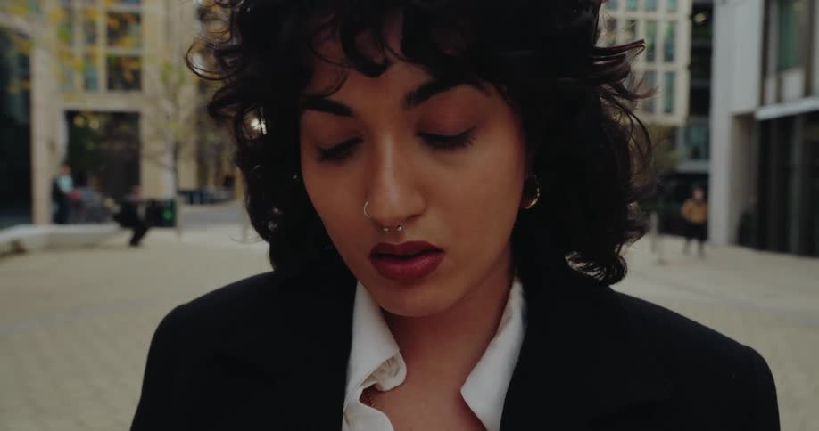 A closeup shot of a woman with curly dark hair wearing a black blazer and white shirt walking outdoors while looking down at her phone she has a nose piercing and dark red lipstickthe background shows a modern city plaza with tall contemporary buil
