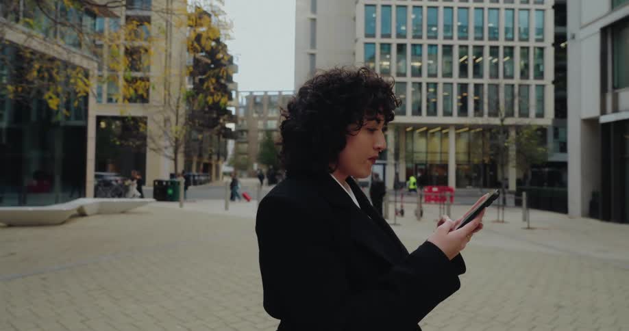 A woman with curly dark hair and a black blazer spin on a paved urban area while looking at her phone