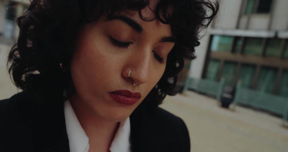 A young woman wearing a suit and a nose ring stands in the middle of the street at midday typing on her phone