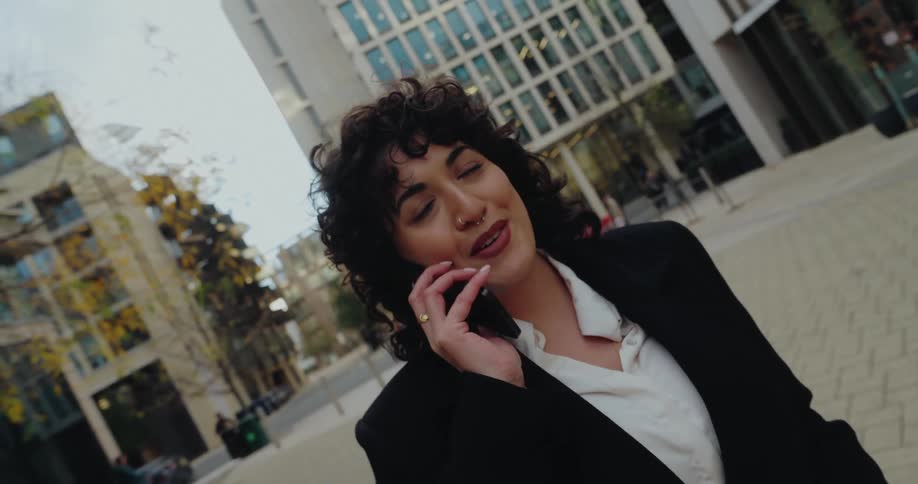 The video begins with a dynamic shot of a woman quickly retrieving a cell phone from her pocket and bringing it to her ear she is walking briskly through an urban area wearing a black blazer and white shirtas she speaks on the phone her expression i