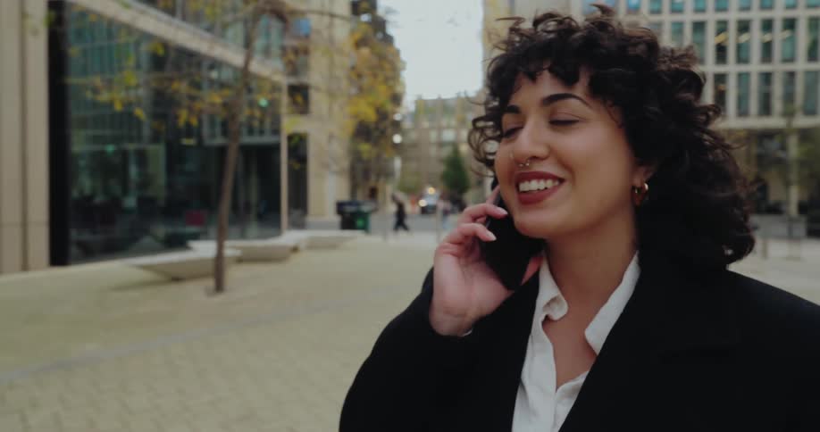 A young woman with curly hair smiles and chats on her phone while walking through a city