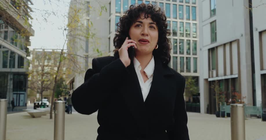 A focused woman in professional black attire is looking forward while talking on a phone in an urban square
