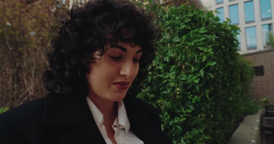 A woman with dark curly hair and a nose ring sits on a stone wall outdoors using her smartphone near green hedges and a modern building