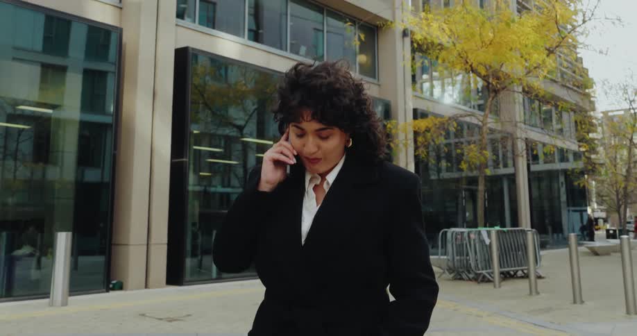 A woman in a black blazer walks along a sidewalk in front of a modern glassfacade building in an urban area speaking intently into her mobile phone while looking forward
