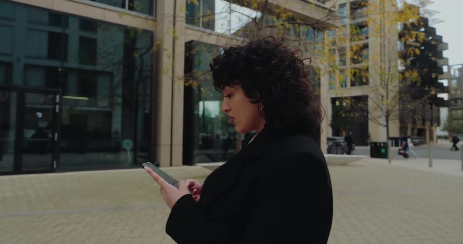 A woman with curly dark hair and a black coat is walking on a brick pavement while focused on using her smartphone near a modern glass building