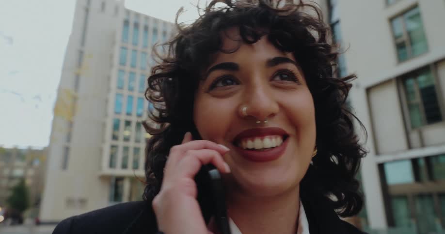 A woman with dark curly hair and a septum piercing wearing a white shirt and black overcoat as she speaks enthusiastically on a mobile phone in a city environment