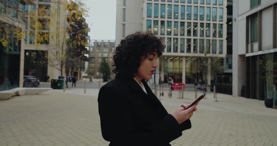 A woman with curly hair stands in a modern city plaza while looking down at her smartphone and texting