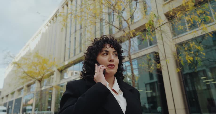 The video features a lowangle shot of a woman with curly hair dressed in professional attire walking outside while talking on a cellphoneshe is situated against the backdrop of a modern building with large glass windows and trees with yellowing aut