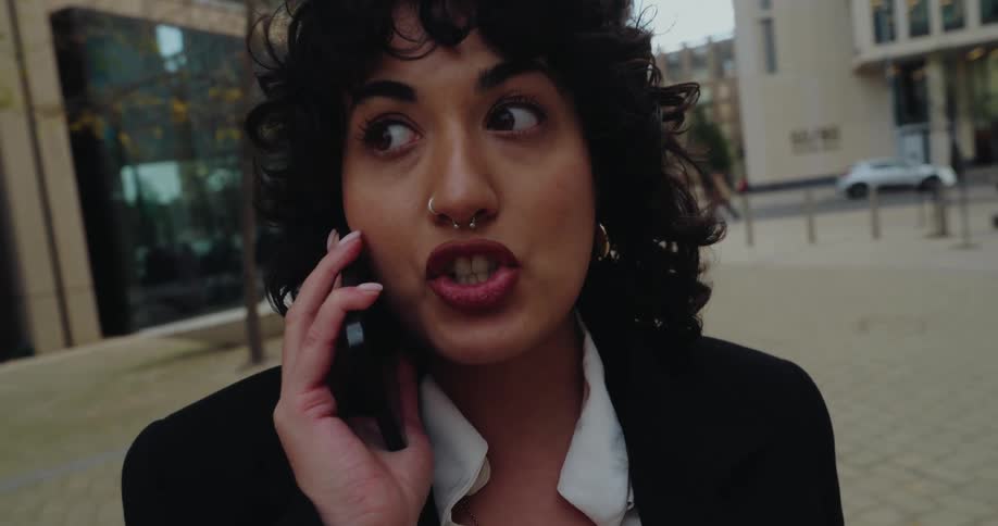 A focused woman in a black blazer and white shirt talks on a cell phone while walking on a paved plaza between modern buildings