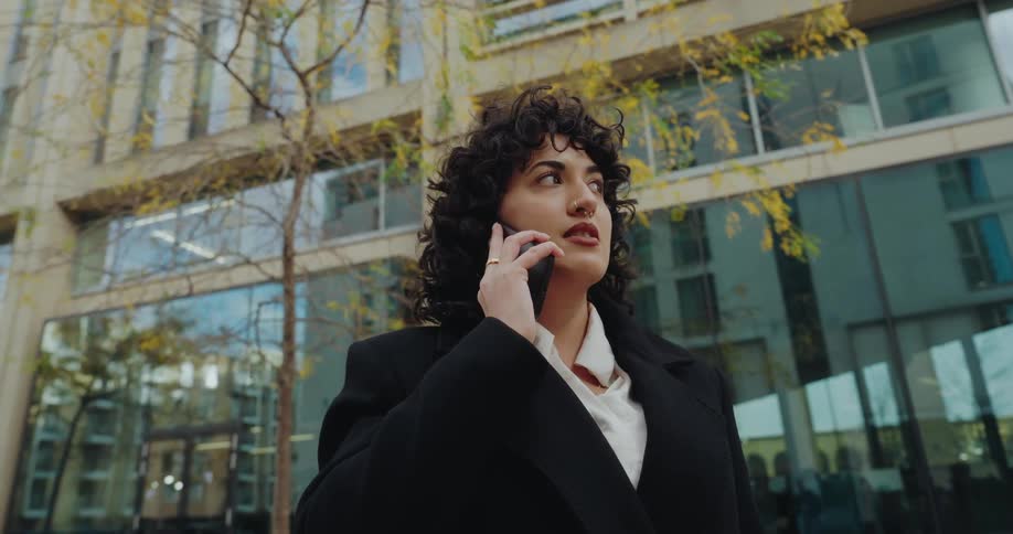 The video features a woman with curly hair dressed in a black coat and white shirt talking on a smartphone outdoors she is walking or standing in an urban setting with modern buildings and trees in the background