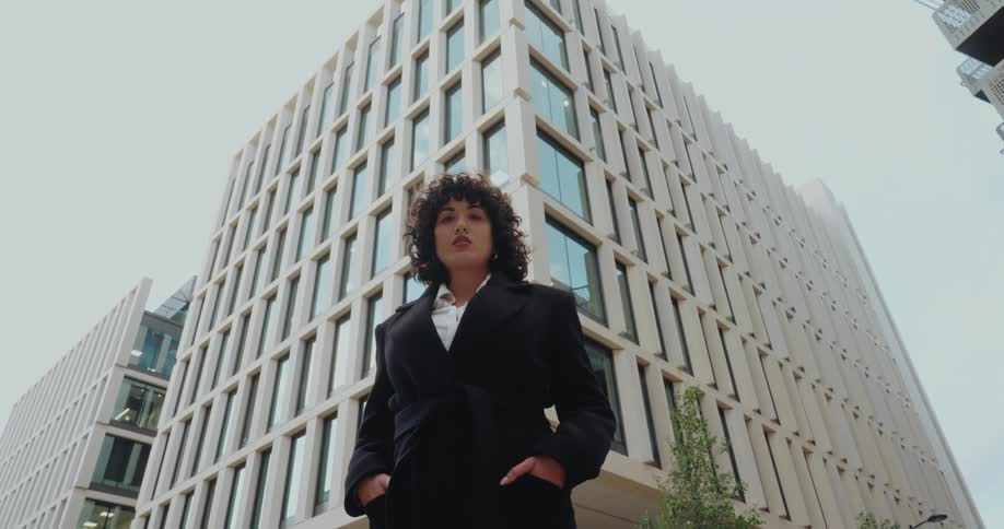 A woman with curly dark hair and a long black coat steps directly toward the camera placing her hands in her pockets and looking up with a direct gaze as the camera captures her from a very low angle emphasizing the modern office building on the back