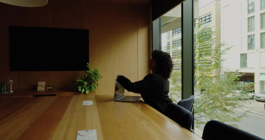 A businesswoman sits at a conference table by a large window and opens her laptop