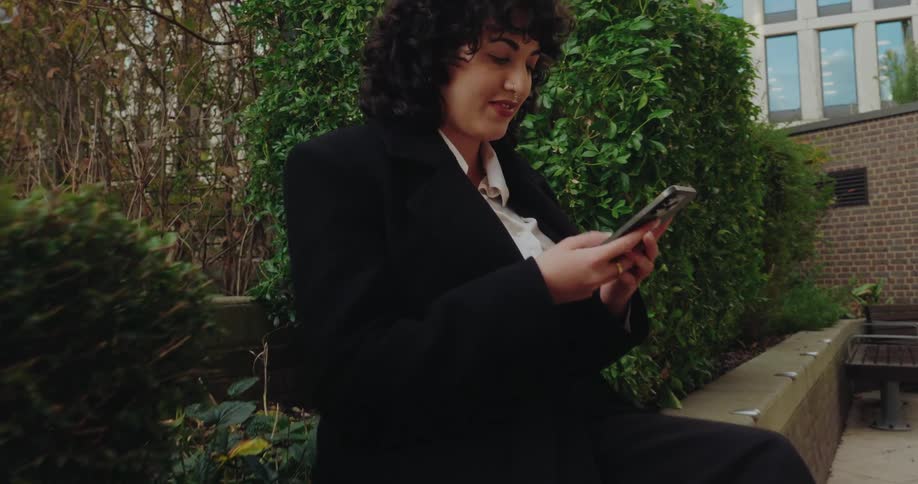 A woman in a black suit and high heels sits on a low brick wall looking at her phone outdoors in an urban green space