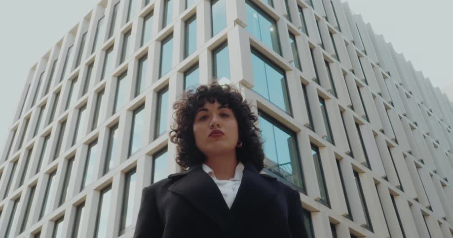This video clip features a lowangle shot of a woman with curly hair wearing a dark coat and looking directly at the camerashe is positioned in front of a modern geometric office building with many vertical rows of windows