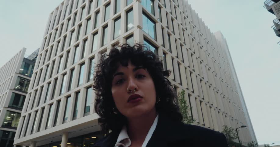 A young woman with black curly hair stands in the middle of a city street at midday wearing a suit and a nose ring