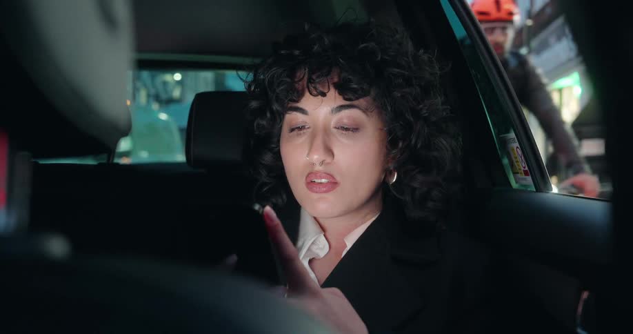 A woman with curly hair is sitting in the back seat of a car at night having a serious conversationshe is gesturing with her hand wearing a black coat and a white shirt with her reflection slightly visible on the screen of a device she is holding of