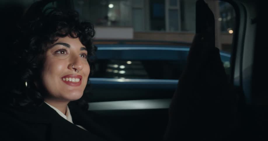 A smiling woman riding in the back seat of a car waves at her smartphone during a video call