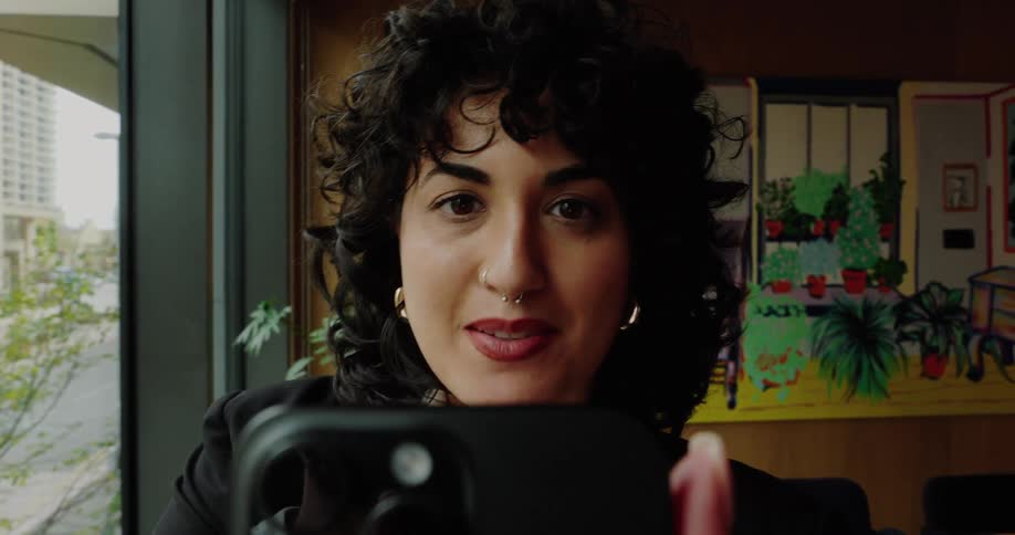 Woman with curly hair and gold hoops holds phone near window with plants in the background