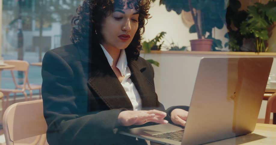A woman in business attire sits at a cafe table working on her laptop