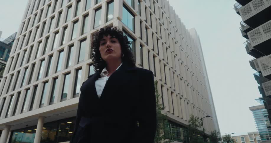 The face of a young woman wearing a black blazer standing in front of a large commercial building and looking ahead