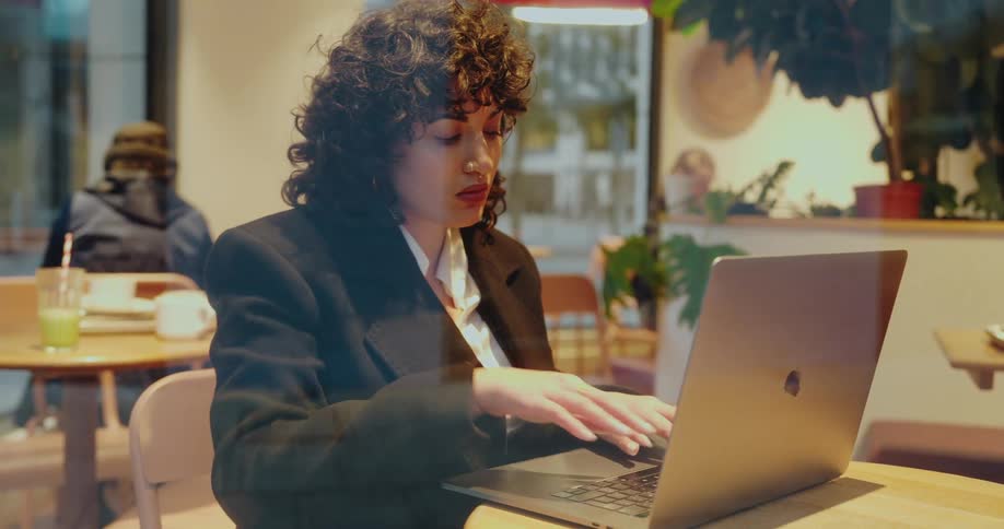 A woman with dark curly hair and a nose ring sits at a wooden table in a brightly lit cafe focused intently on typing on a laptop computer while wearing a black blazer over a white shirt