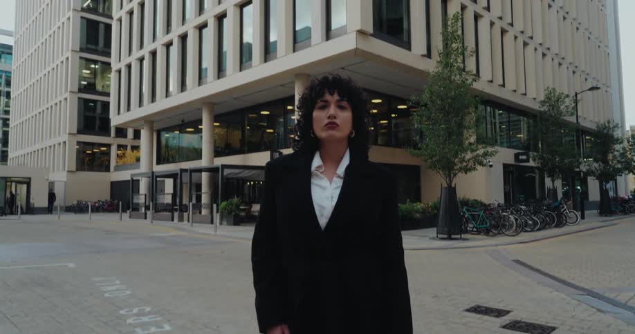This video shows a woman formally dressed in a long black coat and white shirt walking toward and stopping in front of the camera the setting is an urban square with a modern glassfacade building dominating the background and bicycles parked nearby