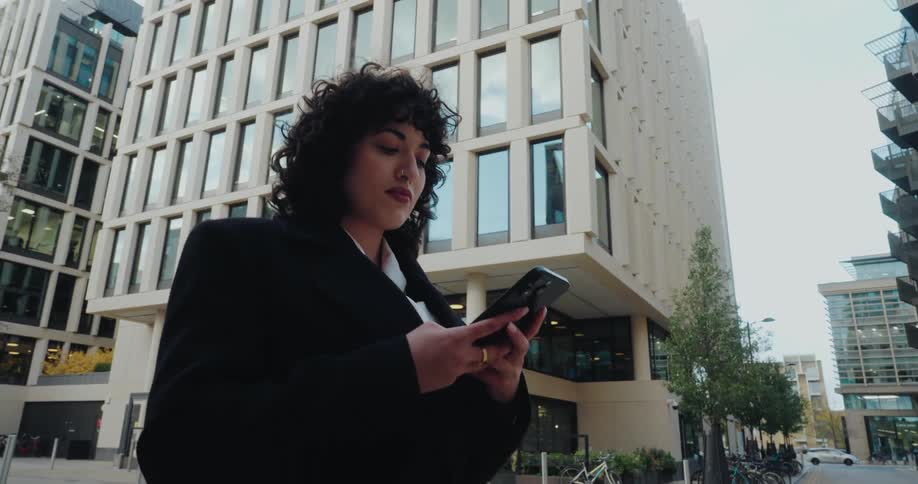 This video shows a lowangle shot of a young woman with curly hair wearing a dark coat standing outside a modern buildingshe is intently focused on her smartphone typing or scrolling on the screen with large office or apartment buildings forming th