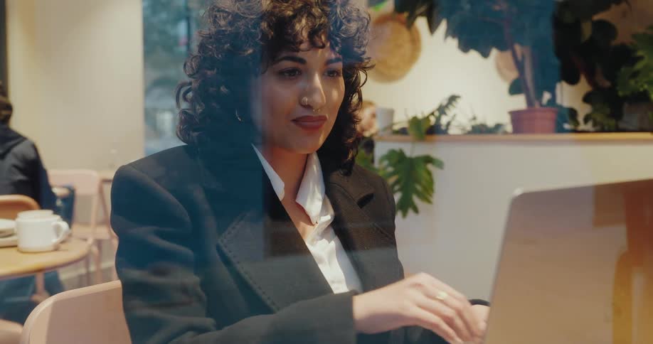 Woman smiles while working on her laptop inside a caf as seen through the window