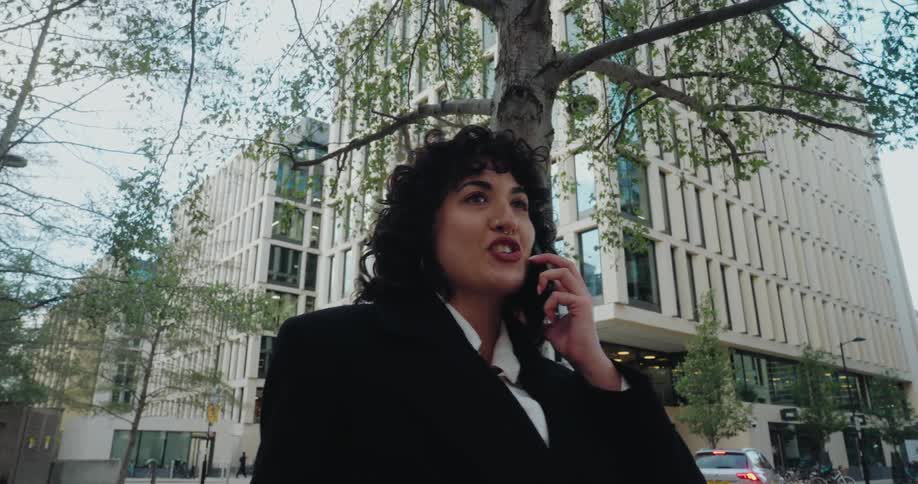 This video clip features a lowangle shot of a woman with curly hair standing outdoors speaking animatedly on a cellphoneshe is dressed in business attire and is framed by the foliage of a tree and a large modern office building in the backgr
