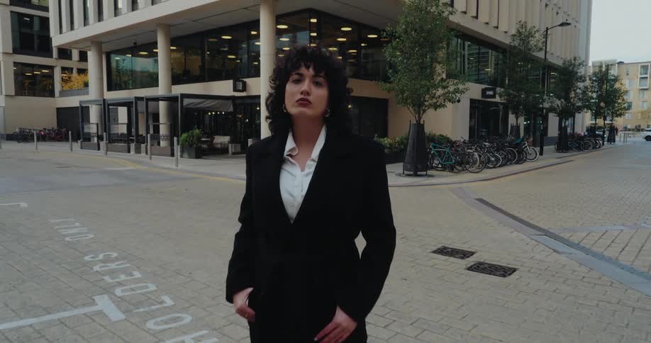 This video shows a woman dressed in a black coat and widelegged trousers over a white shirt walking forward on a paved street and stopping to look directly at the camera the setting is a modern urban square or street corner with a large contemporary
