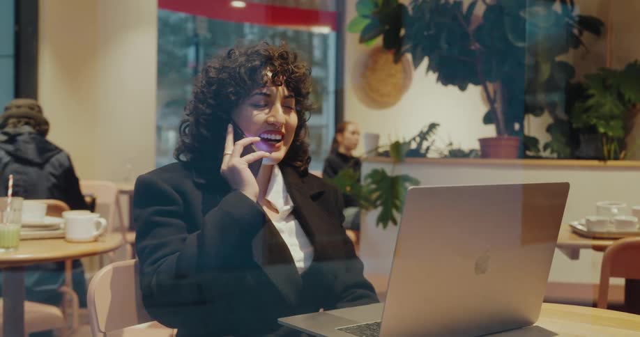 This video captures a professionallooking woman with curly hair working on her laptop in a caf or coworking space she receives a call on her smartphone and smiles suggesting a positive and engaging business or personal conversation while multitasking