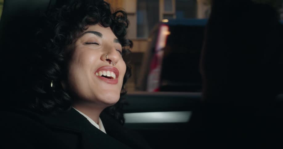 A smiling woman with dark curly hair and a nose ring talks to someone off camera while sitting in a car at night