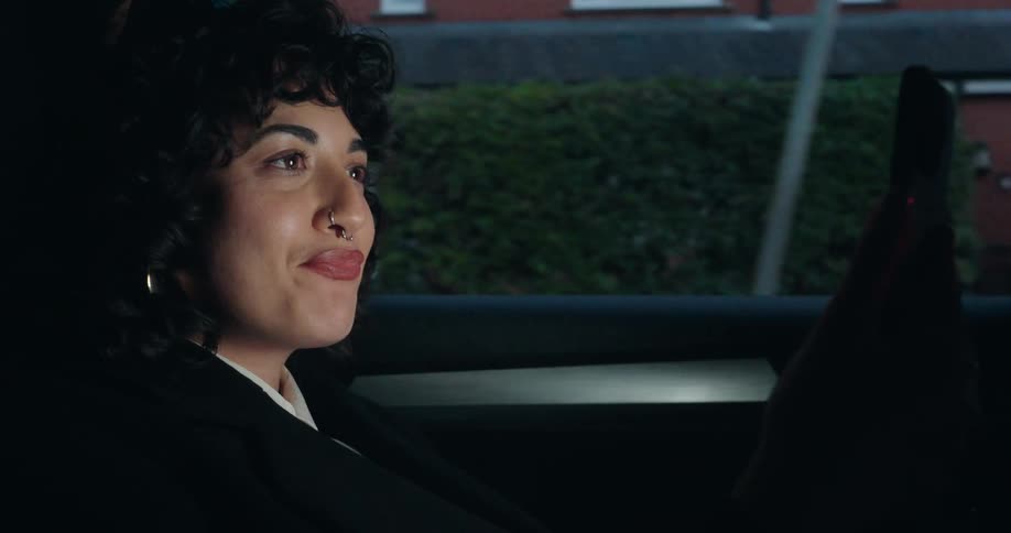 This video features a closeup of a young woman with curly dark hair and a nose ring sitting in the back seat of a carshe is smiling and then begins to speak while holding a phone in her raised hand as the background outside the window shows a reside