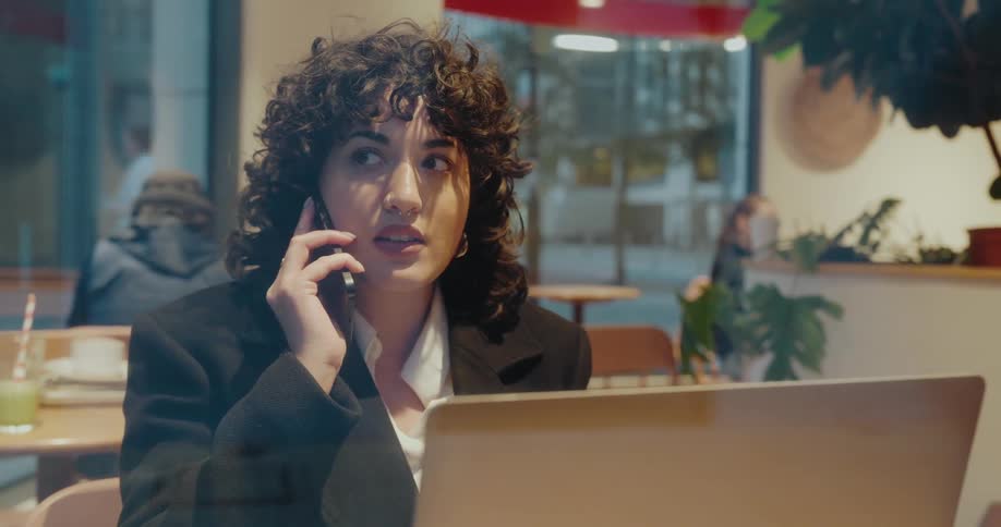 Woman sits at a table with a laptop computer as she talks on her phone