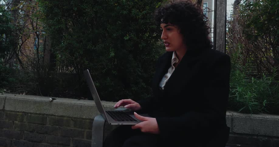 A woman with dark curly hair professionally dressed in a black blazer and white shirt sits outdoors on a bench with a laptop on her lap alternately smiling and speaking seriously