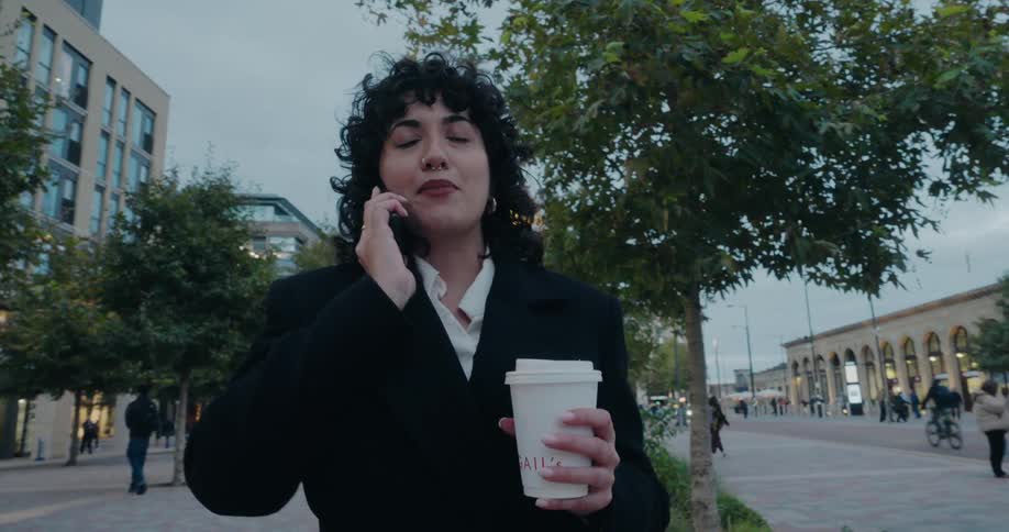 This video shows a young woman having a conversation on a mobile phone while standing on a city street or plazashe has dark curly hair and is wearing a dark jacket or coat over a white collared shirt she is holding a white takeaway cup that appears