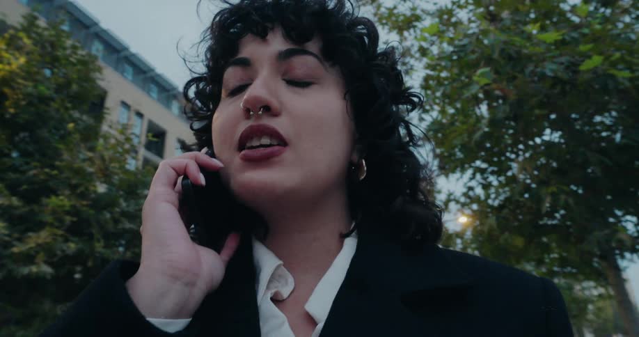 Woman standing in an urban plaza holds a to go cup while talking on a smartphone in the evening