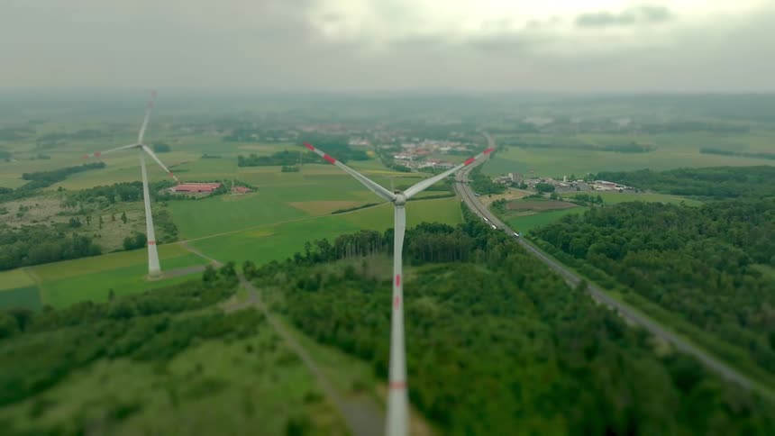 Two wind turbines are looking forward over a rural landscape with a highway and small town under a cloudy sky