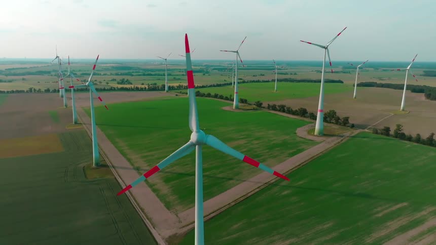 An aerial view of a wind farm with multiple white turbines with red accents standing in green agricultural fields under a bright hazy sky