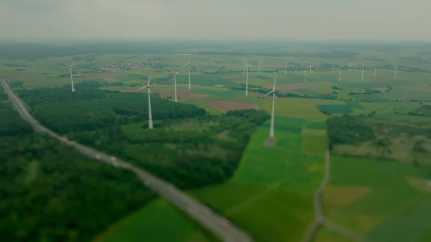 A highangle aerial view captures a large wind farm consisting of numerous white turbines spread across a wide flat landscape of green fields and small forests bisected by a busy highway in the foreground under an overcast sky
