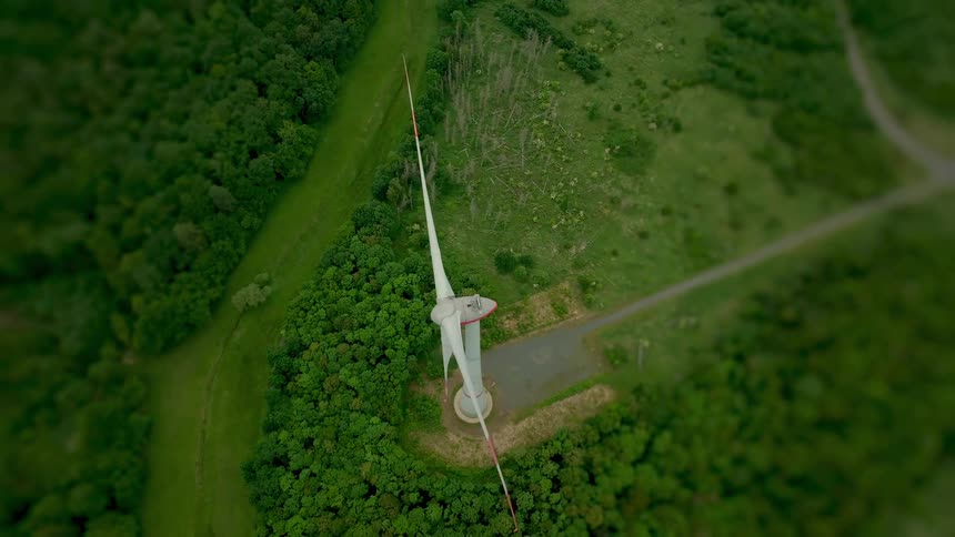 An aerial topdown shot descends directly over a large threebladed wind turbine which is actively rotating and stands on a clearing within a dense primarily green forested and natural landscape