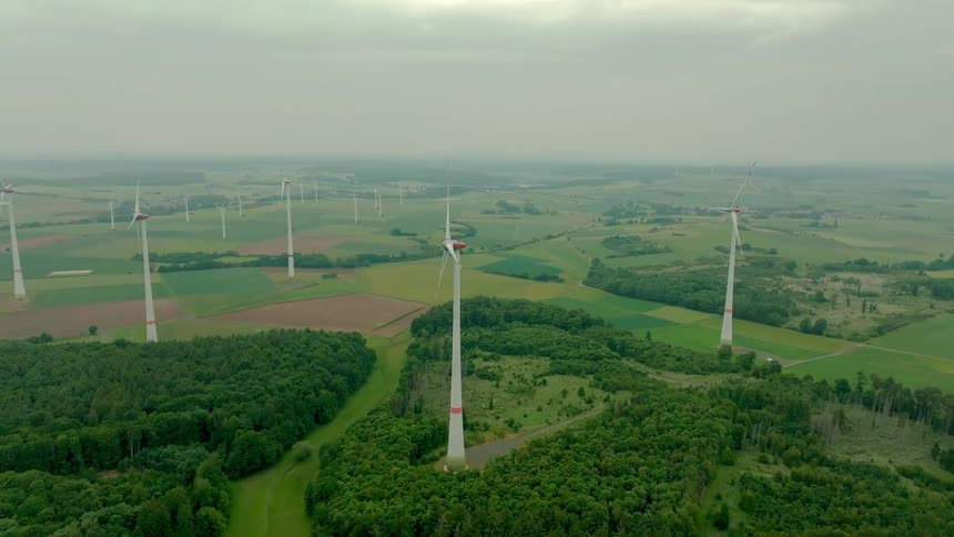 An aerial view of a wind farm with turbines across forests and green fields under an overcast sky