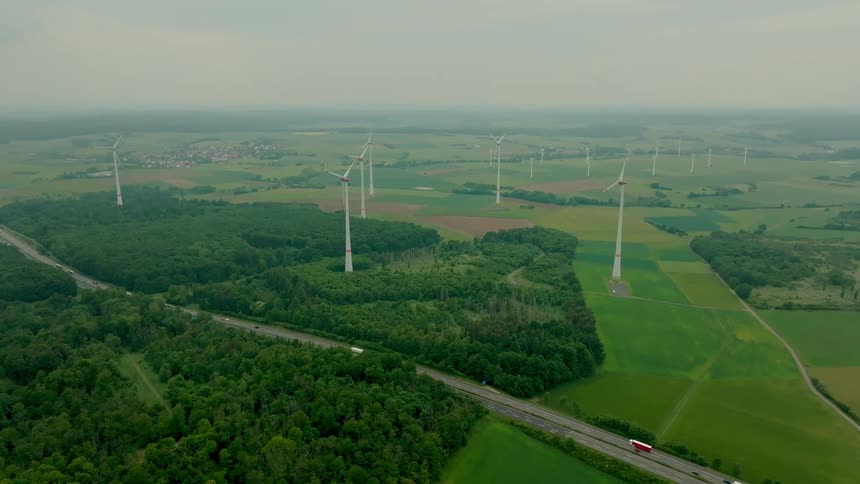 Aerial view of wind turbines between a highway and farmland under an overcast sky