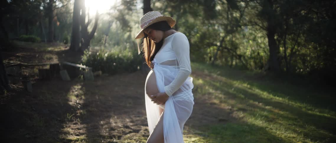 Pregnant woman in a white dress cradles her belly in a sunlit clearing