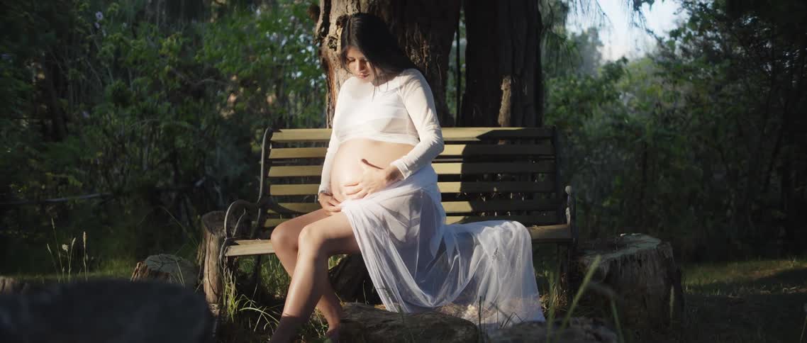 A pregnant woman sits on a bench embracing her baby bump in a park