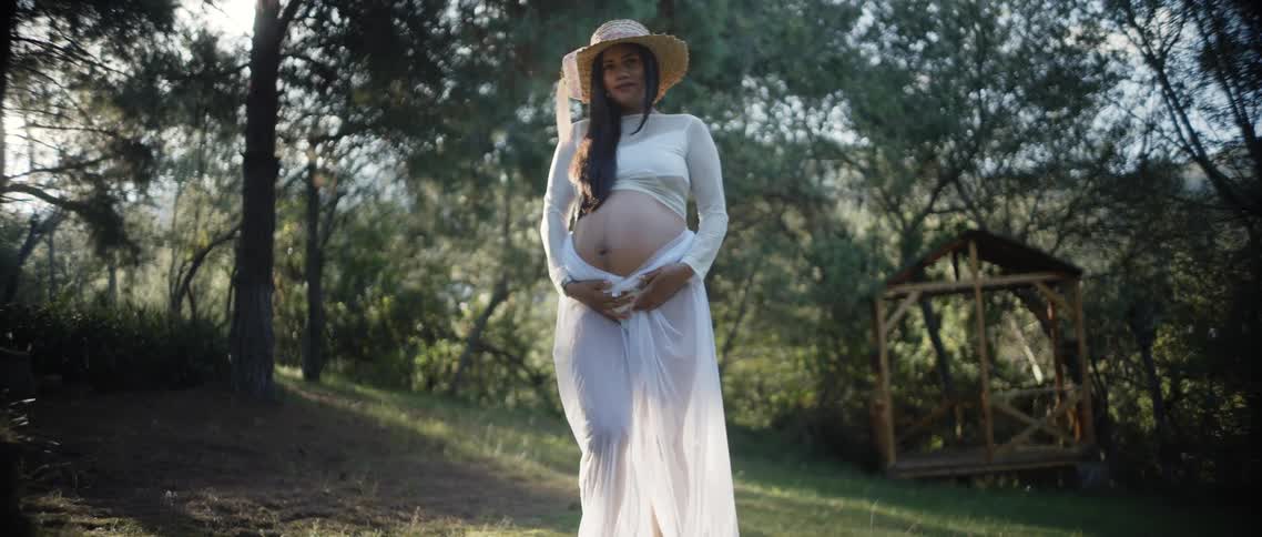 Pregnant woman poses outdoors holding her belly among sunlit trees