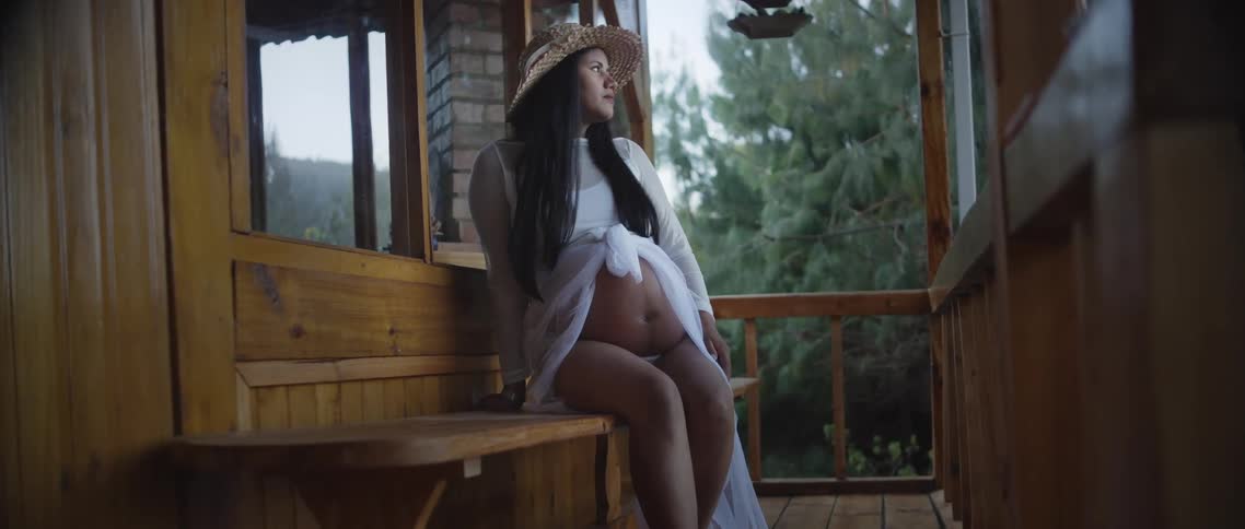 The video features a pregnant woman with long dark hair sitting on a wooden bench on a rustic balcony or porch overlooking a dense lush green landscape of trees she is wearing a straw hat and is minimally covered in a sheer white wrap exposing her ab