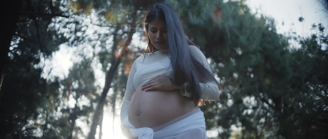 A pregnant woman stands in a sunlit forest gently cradling her belly with a peaceful expression