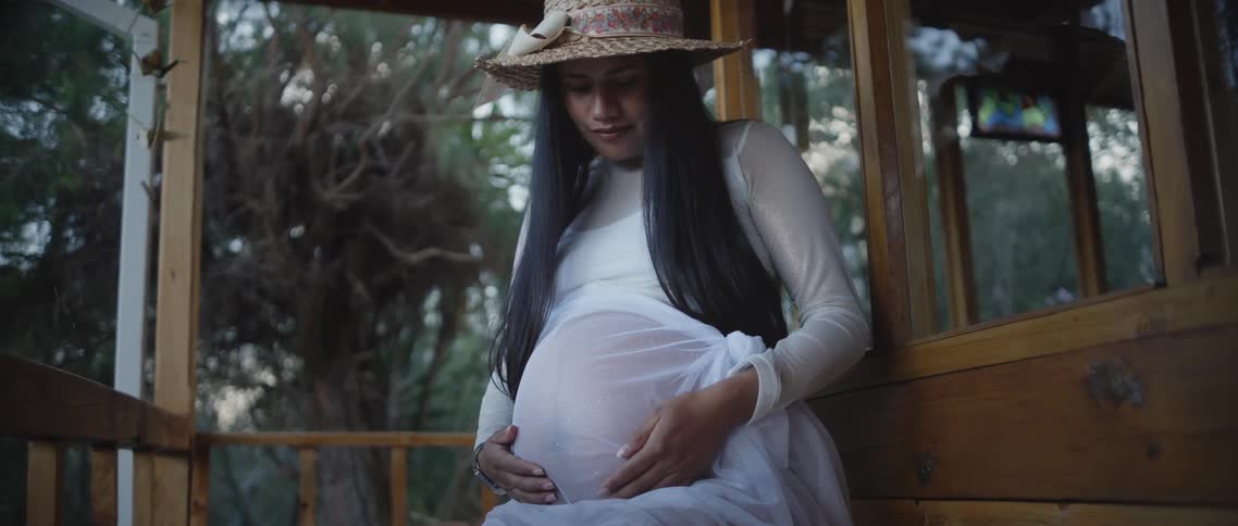 Happy woman holding her pregnant belly sitting outdoors on porch in the forest